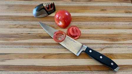 overhead view of Teakhaus Traditional Edge Grain Professional Cutting Board with Henckels knife, partially sliced tomato, and a KitchenIQ manual knife sharpener.